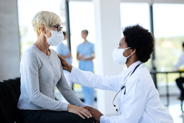 검은 의사와 성숙한 환자는 병원에서 대기실에서 이야기하는 동안 보호 얼굴 마스크를 착용. - doctor and patient 뉴스 사진 이미지
