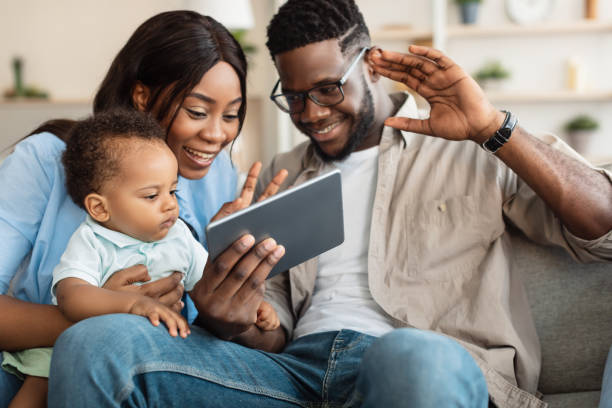Black family having video call using tablet waving hands Family, Parenthood and Technology Concept. Portrait of happy smiling black parents holding little baby on hands having video call on tablet computer at home, waving to screen, talking with relatives black child tablet stock pictures, royalty-free photos & images