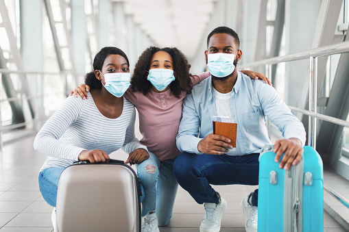Travel Safe During Coronavirus Pandemic. Portrait of African American family of three wearing protective medical masks posing, sitting with passports, tickets and luggage at airport terminal