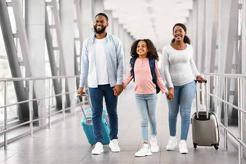 Family Travel Concept. Portrait of excited black dad, mum and girl walking at airport terminal corridor with suitcases waiting for the aircraft arrival, three people holding hands, free copy space