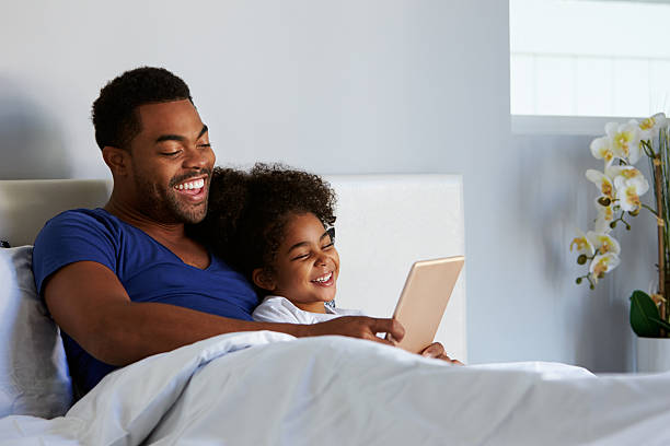 Black father and daughter relax in bed with tablet computer
