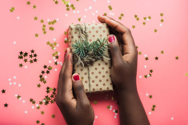 black girl is holding a gift of kraft paper on a pink background with confetti. womans hands holding gift or present box decorated confetti on pink pastel table top view. flat lay composition for birthday or wedding or christmas or happy new year - happy birthday celebrity imagens e fotografias de stock