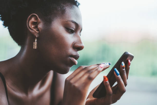 Black girl is using a smartphone The portrait of a charming young black female with multi-colored nail-art using a smartphone while sitting on the summer street; an exquisite African girl with earrings and the cellphone outdoors beautiful polish girls stock pictures, royalty-free photos & images