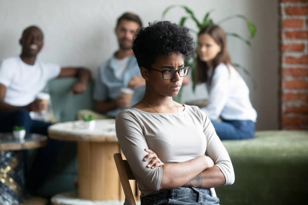 Black girl outcast sitting apart from peers in cafeteria Unkind teenagers looking laughing at black classmate, focus on mixed race unhappy shy and frustrated girl wearing glasses sitting at closed posture arms crossed feeling insecure having low self-esteem judgement stock pictures, royalty-free photos & images
