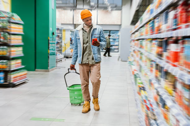 Black guy with basket and phone walks along supermarket stock photo