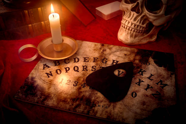 black magic and contacting the dead concept A witches den, fortune teller and black magic concept with a talking spirit board lit by a candle surrounded by a deck of cards, three books and a skull engulfed in mysterious and murky smoke ouija board stock pictures, royalty-free photos & images
