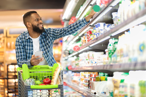 Black Male Buyer Shopping Groceries In Supermarket Taking Dairy Product From Shelf Standing With Shop Cart Indoors. Guy Buys Grocery Choosing Food In Super Market. Empty Space