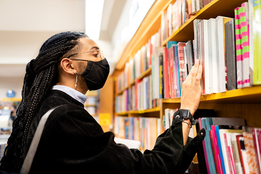 University student, at a library choosing a book in the shelf, using facial mask