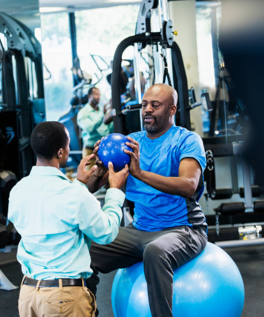 A physical therapist working with a patient in the gym, helping the man recover from an injury by doing strengthening exercises. They are both African-American men. The patient, in his 50s, is sitting on a fitness ball and the PT is kneeling in front of him. They are passing a medicine ball back and forth.