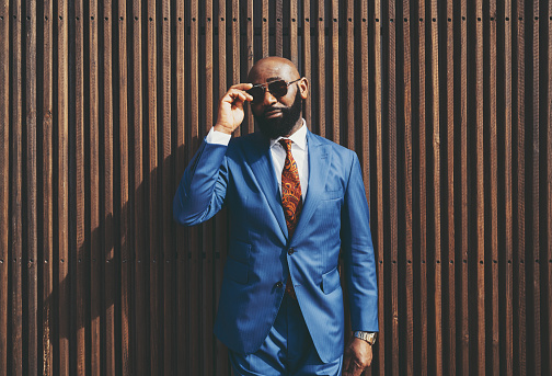 The portrait of a burly handsome bald black guy with a well-trimmed beard, in an elegant custom blue formal suit with a necktie, adjusting sunglasses outdoors in front of a wooden striped background
