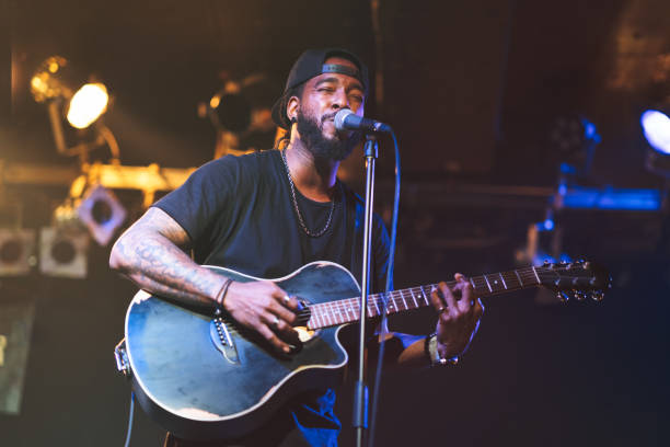 Black man playing acoustic guitar and singing on stage A black man is playing the acoustic guitar and singing passionately on stage. singer stock pictures, royalty-free photos & images