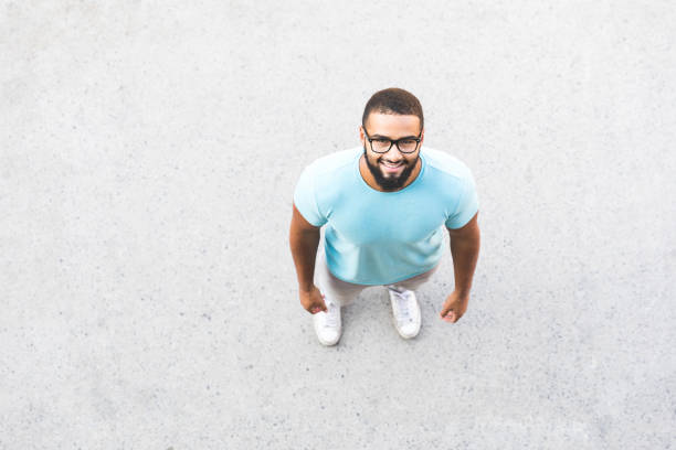 Black man portrait. Over head shot Black man portrait. Over head shot, the man is looking up at camera. He is wearing black eyeglasses and a light blue t-shirt. Smiling man on a concrete pavement directly below stock pictures, royalty-free photos & images