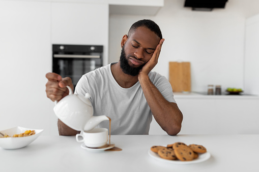Lack Of Sleep Concept. Portrait of tired young African American man sleeping while sitting at dining table in kitchen, holding kettle pouring coffee away from cup spilling hot drink on desk