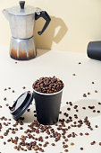 istock Black paper coffee cup is filled with coffee beans. In the background there is a coffee maker and an empty coffee glass. 1339272362