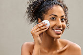 Young pretty african american woman taking off her makeup with cotton wipe sponge. Smiling girl cleaning face with cotton pad isolated over background. Black young woman cleansing face, daily healthy beauty routine.