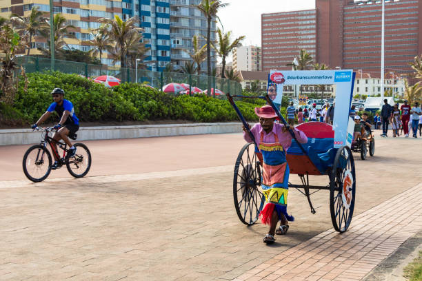 A black south african man carrying a rickshaw without passengers at the beach front of Durban, South Africa. Durban, South Africa - January 6th, 2019: A black south african man carrying a rickshaw without passengers at the beach front of Durban, South Africa. durban stock pictures, royalty-free photos & images