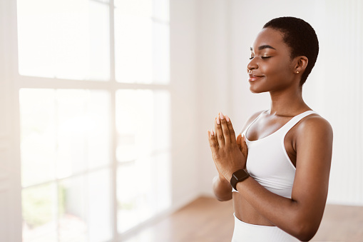Serenity And Mindfulness Concept. Portrait of calm smiling African American female holding hands in prayer pose, keeping palms together, meditating, practicing Kundalini Yoga. Free copy space