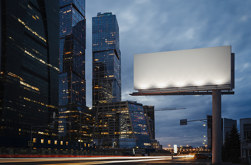 Blank billboard at night time in the city next to skyscrapers and road with lights on the frame. 3d rendering