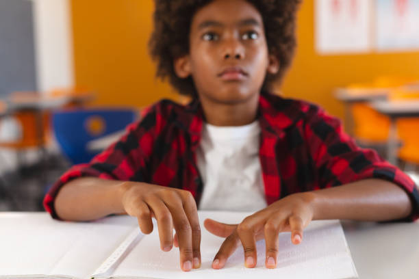 Blind african american schoolboy sitting at desk in classroom reading braille book with fingers stock photo