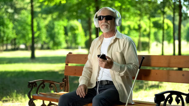 anciano ciego usando auriculares escuchando audiolibro, mensaje de voz en el teléfono celular - ceguera fotografías e imágenes de stock