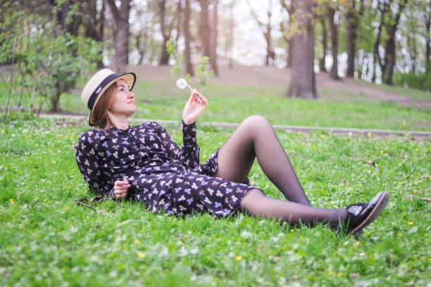 blonde girl in a straw hat with a dandelion in his hand lies  on the grass in the spring garden. Spring season concept. stock photo