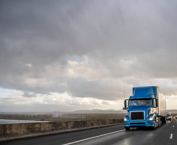 Blue big rig semi truck transporting cargo in dry van semi trailer running on the wet highway road with stormy rain sky stock photo