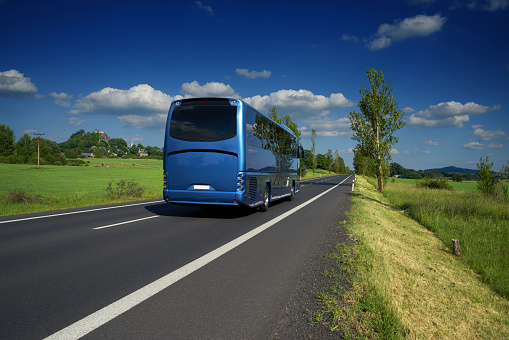 Blue bus traveling on asphalt road in a rural landscape. Village and mountain with castle ruins in the background.