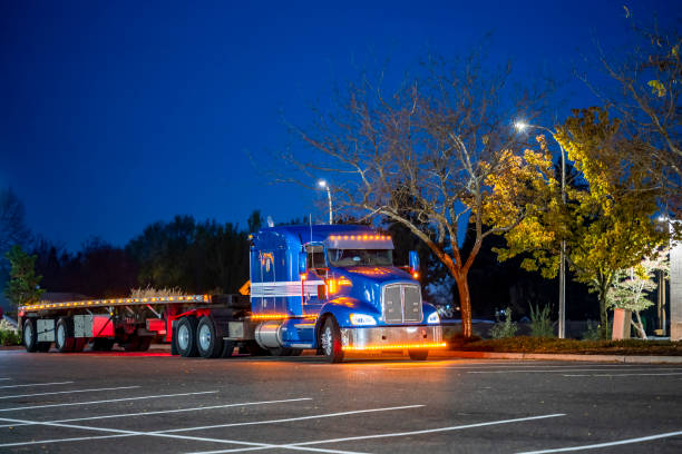Blue classic big rig semi truck with turned on lights and flat bed semi trailer rest standing on the truck stop parking lot at night time stock photo