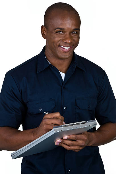 Blue collar worker writing African American mechanic writing on clipboard and looking up with a pleasant smile  african american plumber stock pictures, royalty-free photos & images