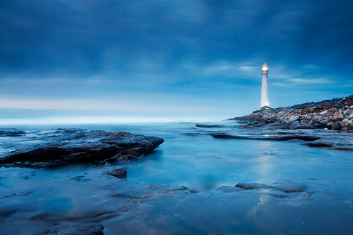 Soft blue hues of early evening, and a subtle reflection of the lighthouse in the water in the foreground. Photographed on a Canon 5D mkiii and an ND Grad filter, with slow shutter speed to create soft, moody water. This is the Slangkop lighthouse in Kommetjie, on the Cape peninsula near Cape Town, South Africa. 