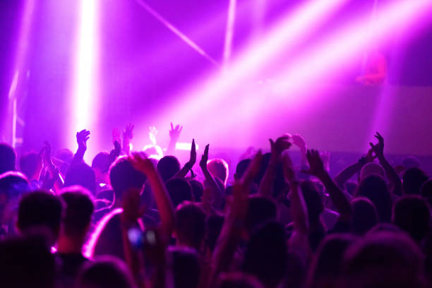 Blurred image of silhouette of raised arms, crowd of people in the front of bright stage lights at concert stock photo