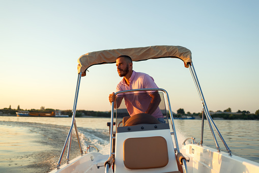 A handsome young man wearing a pink shirt is steering a motorboat at sunset on the Danube river.