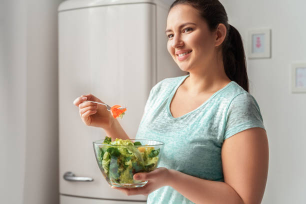 Body Care. Chubby girl standing in kitchen eating healthy salad smiling cheerful Young chubby woman standing in kitchen eating healthy fresh green salad looking aside smiling cheerful fat nutrient stock pictures, royalty-free photos & images
