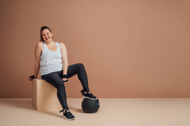 Body Positive Fitness: Studio Portrait of Plus Size Woman Model in Sportswear (Copy Space) Pretty Caucasian smiling plus size woman model sitting by medicine ball in sportswear and looking at camera. beautiful voluptuous women stock pictures, royalty-free photos & images