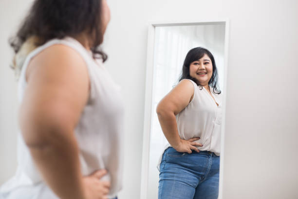 Body positive plus size female clothing. Body positive plus size woman happily and is proud of herself looking at mirror at home. Try wearing clothes that measure the size. stock photo