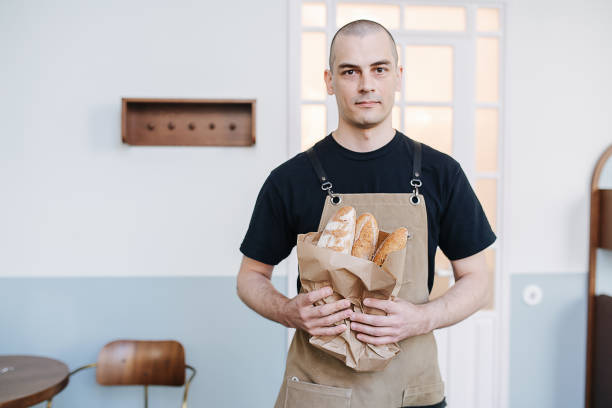 Bold male baker holding paper bag with fresh baguettes stock photo