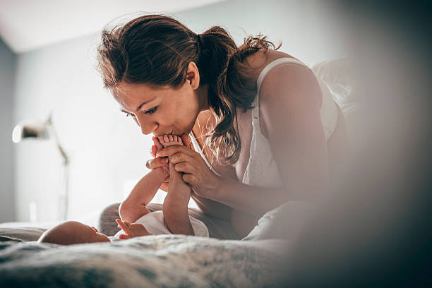 Bonding with my baby A happy new mother smiles as she kisses her newborn's feet while relaxing on her bed. The baby is unrecognisable as she lies down looking up at her mother. human toe stock pictures, royalty-free photos & images