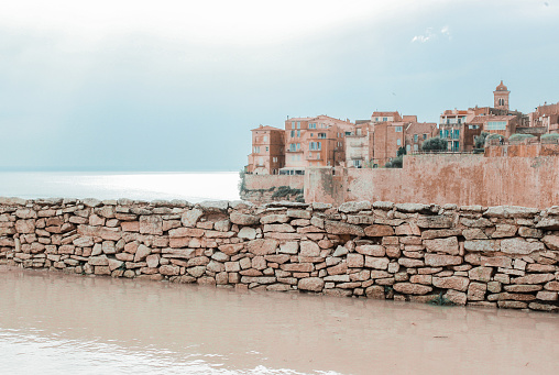 Old pink, orange houses on rocky mountain right by dark blue sea. The facades are bricked and ancient. Cloudy, rainy sky with clouds breaking through which the sun shines. Light colors. In the foreground is an old wall and in front of it is a large puddle of water due to the rain.