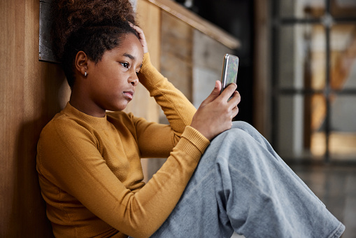 Sad African American girl text messaging on smart phone while relaxing at home.