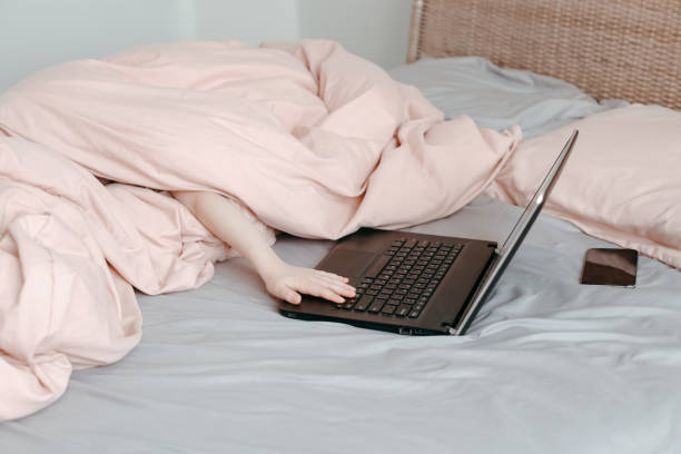 скучный забавный ребенок, лежащий в постели под одеялом и учающийся в виртуальном онлайн школьном классе. руки, печатающие на клавиатуре ко - blue monday стоковые фото и изображения