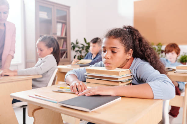 Bored mixed-race schoolgirl in casualwear keeping chin on top of stack of books Bored mixed-race schoolgirl in casualwear keeping her chin on top of stack of books by desk on background of classmates and teacher at lesson students stock pictures, royalty-free photos & images