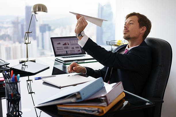 Bored White Collar Worker Throwing Paper Airplane In Office Corporate manager in modern office takes a break and prepares a paper airplane. The bored man dreams of his next vacations and leans back on his chair wasting time stock pictures, royalty-free photos & images