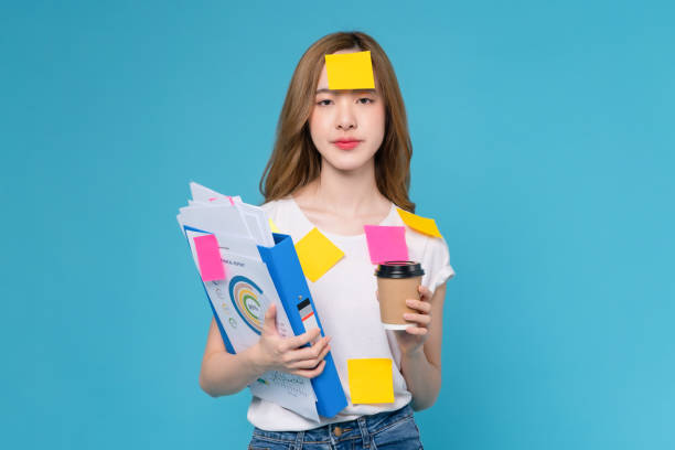 Boring young Asian woman holding document file with coffee cup and a sticky note on the face, stand on studio shot blue background. stock photo
