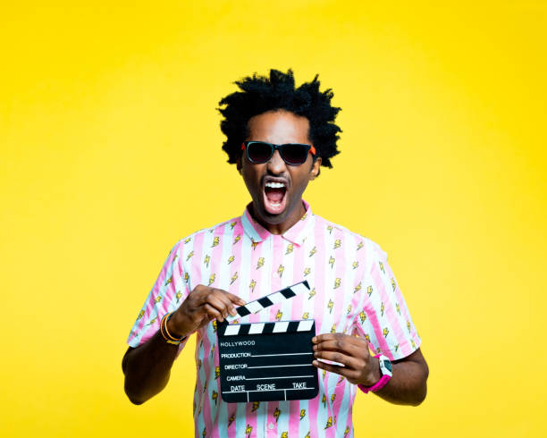 Potret musim panas pemuda afro amerika yang marah dengan rambut gimbal mengenakan kemeja vintage dengan pola kilat dan kacamata hitam, memegang papan tulis papan tulis film, berteriak ke kamera. Studio ditembak dengan latar belakang kuning.