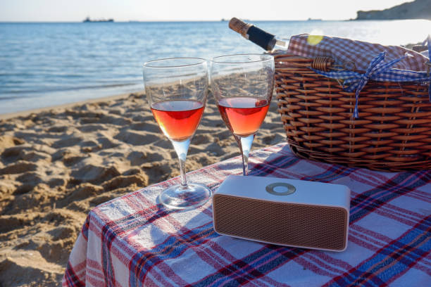A bottle of rose wine in a wicker picnic basket, a bluetooth speaker and two glasses of rose wine next to it on a plaid tablecloth. A bottle of rose wine in a wicker picnic basket, a bluetooth speaker and two glasses of rose wine next to it on a plaid tablecloth. Focus on foreground. A romantic summer vacation concept. bluetooth stock pictures, royalty-free photos & images