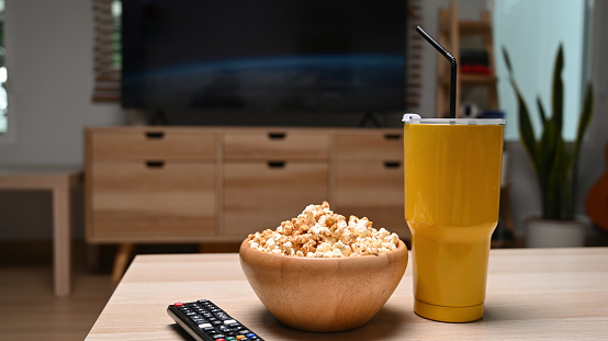 Bowl of popcorn, remote control and glass of beverage on wooden table.