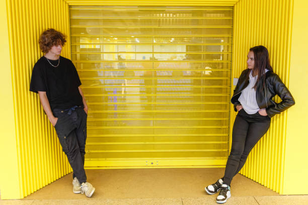 A boy and a girl leaning against a yellow wall looking at each other with their hands in their trouser pockets. stock photo
