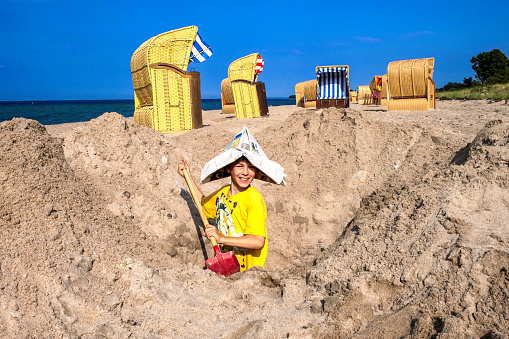 Boy digs a deep hole with a shovel in the sand on the beach by the sea