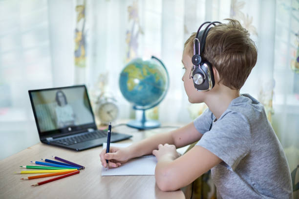 A boy in headphones looks at a laptop while doing homework at home stock photo