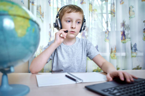 Boy in headphones talking through a laptop while doing homework at home stock photo
